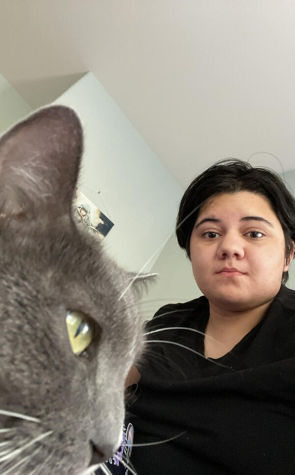 lani, a masc person with a round face and short black hair, standing next to Lola, a grey cat